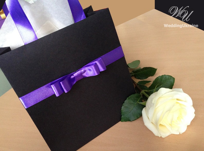 May include: A black gift bag with a purple ribbon and bow. A single white rose with green leaves is next to the bag.