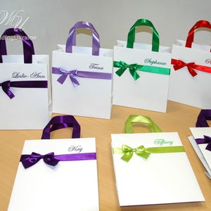 May include: Six white gift bags with different colored ribbon bows and personalized names. The names are: Leslie-Ann, Trina, Stephanie, Robyn, Kay, and Tiffany.