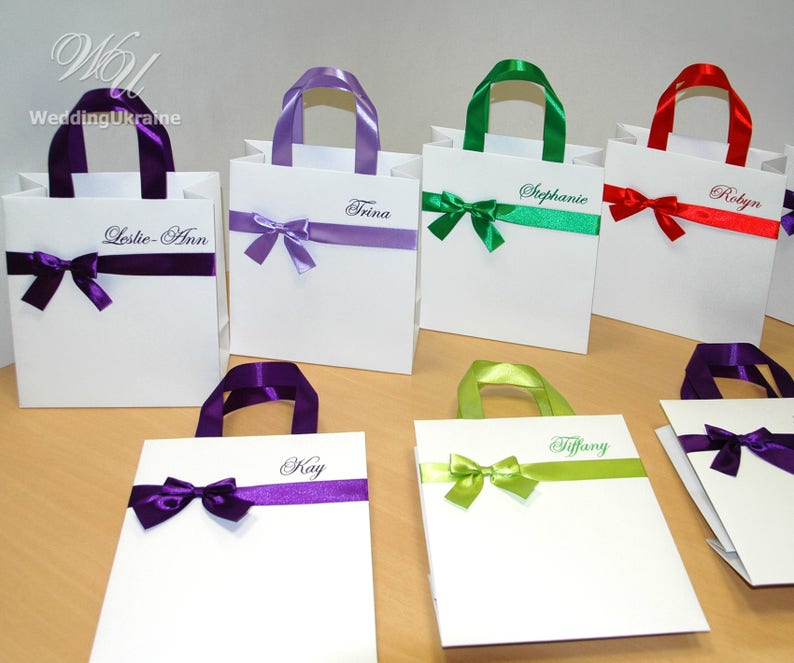 May include: Six white gift bags with different colored ribbon bows and personalized names. The names are: Leslie-Ann, Trina, Stephanie, Robyn, Kay, and Tiffany.