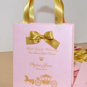 May include: Pink gift bag with gold ribbon and bow. The bag has a gold crown and the text "Thank You for Welcoming Our Sweet Little Princess Skylar Jhene October 2019" printed on it. The bag also has a gold horse and carriage illustration.