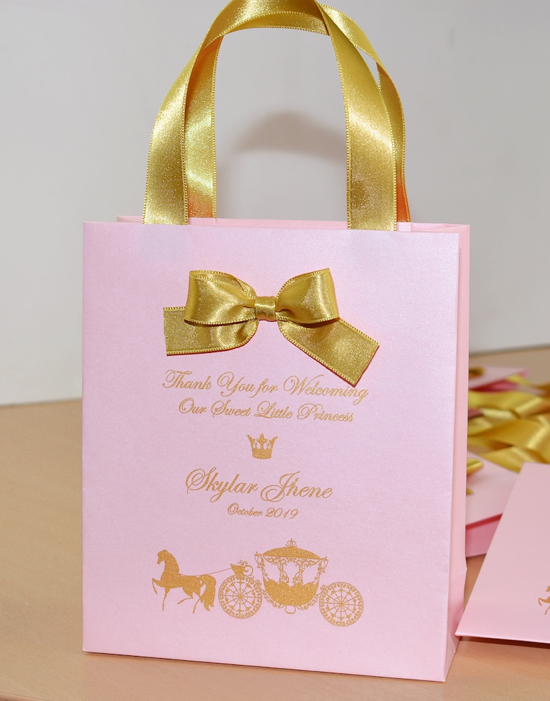 May include: Pink gift bag with gold ribbon and bow. The bag has a gold crown and the text "Thank You for Welcoming Our Sweet Little Princess Skylar Jhene October 2019" printed on it. The bag also has a gold horse and carriage illustration.