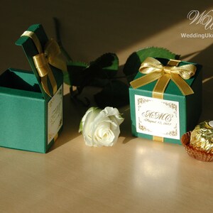 May include: Two green favor boxes with gold ribbon and a personalized label. The label says "A.M.C. August 12, 2017". A chocolate candy is in the background.