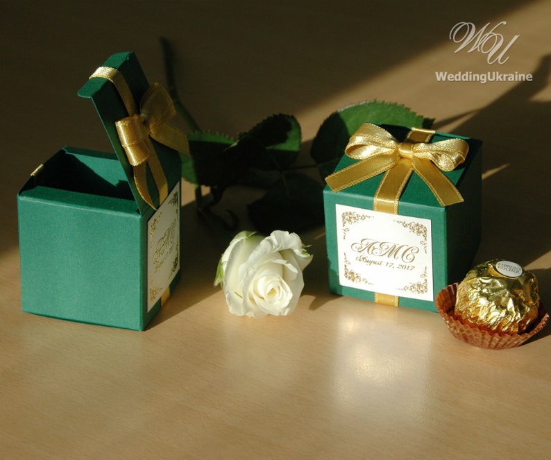 May include: Two green favor boxes with gold ribbon and a personalized label. The label says "A.M.C. August 12, 2017". A chocolate candy is in the background.