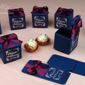 May include: Dark blue gift boxes tied with burgundy ribbon bows. Each box has a gold label with the names "Natasha & Dennis" and a date. Two gold-wrapped chocolates are also visible. Wedding favours.