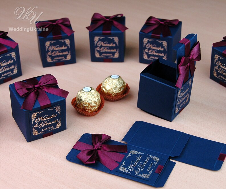 May include: Dark blue gift boxes tied with burgundy ribbon bows. Each box has a gold label with the names "Natasha & Dennis" and a date. Two gold-wrapped chocolates are also visible. Wedding favours.