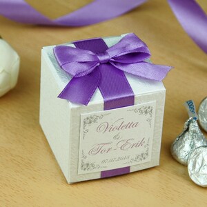 May include: White favor box with a purple satin ribbon and bow. The box has a label with the text "Violetta & Tor-Erik" and the date "05.07.2018".
