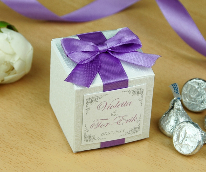 May include: White favor box with a purple satin ribbon and bow. The box has a label with the text "Violetta & Tor-Erik" and the date "05.07.2018".
