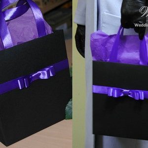 May include: A black gift bag with purple ribbon and bow. The bag is filled with purple tissue paper. The bag has a purple handle.