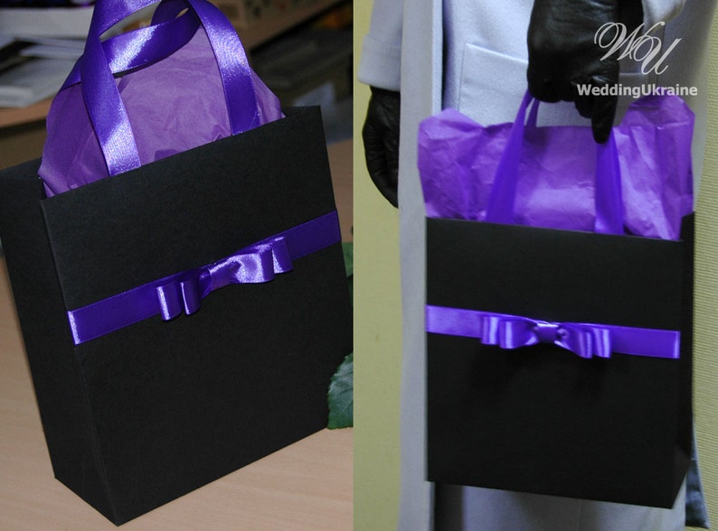 May include: A black gift bag with purple ribbon and bow. The bag is filled with purple tissue paper. The bag has a purple handle.