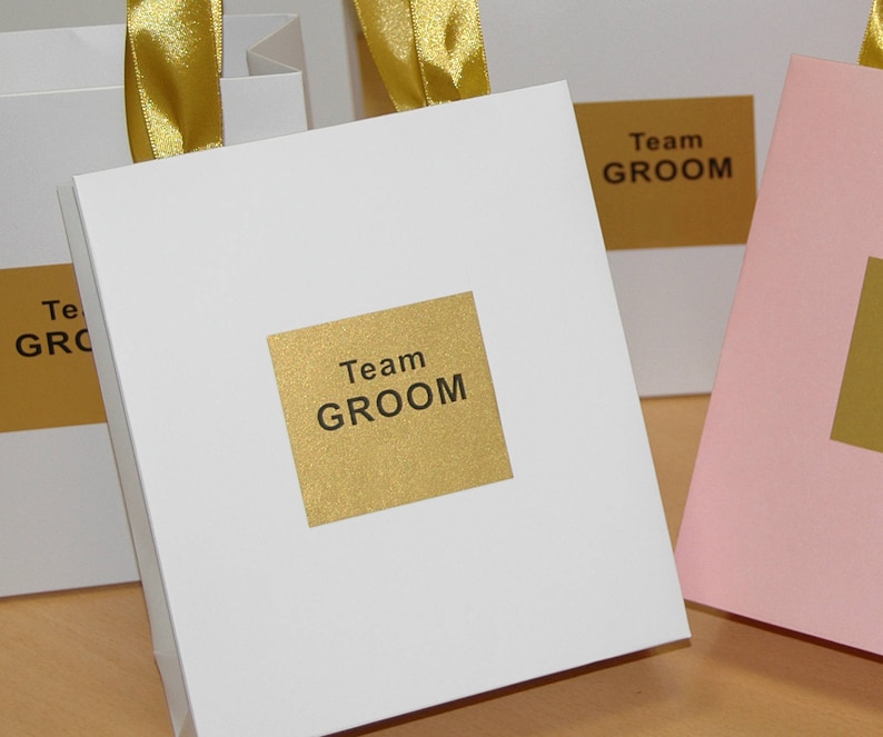 May include: White gift bags with gold ribbon and gold labels that say "Team Groom".