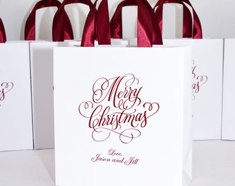 May include: White gift bags with red satin handles. The bags have a red cursive inscription that says "Merry Christmas" with a heart and the text "Love, Jason and Jill".