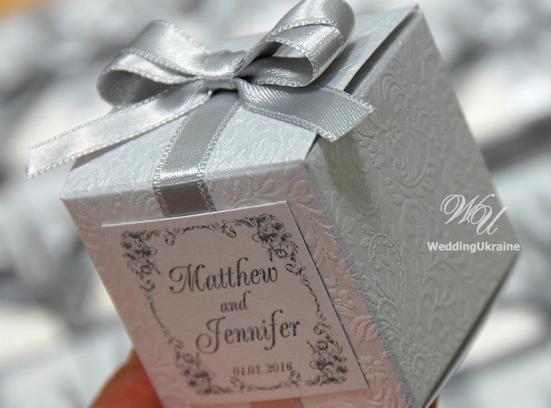 May include: A white favor box with a silver satin ribbon and bow. The box has a decorative embossed pattern and a label that reads "Matthew and Jennifer 01.01.2018".