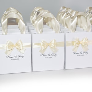Puede incluir: Bolsas de regalo blancas con lazos de cinta de sat&eacute;n marfil y texto personalizado. Las bolsas dicen "Kevin & Patty 10.03.2020".