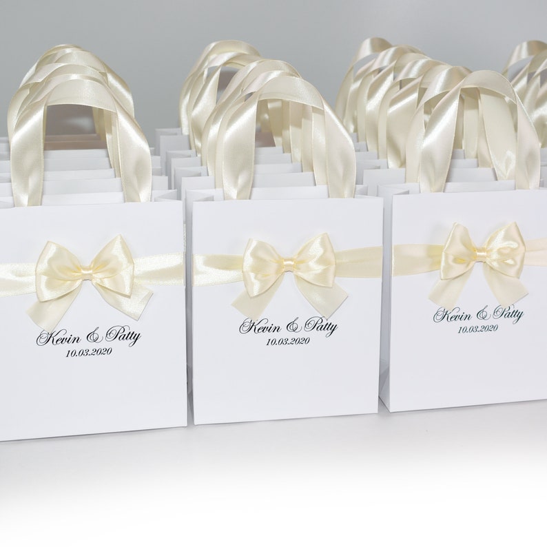 Puede incluir: Bolsas de regalo blancas con lazos de cinta de sat&eacute;n marfil y texto personalizado. Las bolsas dicen "Kevin & Patty 10.03.2020".