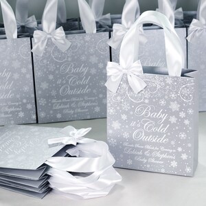 May include: Silver gift bags with white satin ribbon bows and snowflake designs. The bags have the text "Baby it's Cold Outside" and "Thanks Snow Much for Coming to Lakiah & Stephen's Baby Shower".
