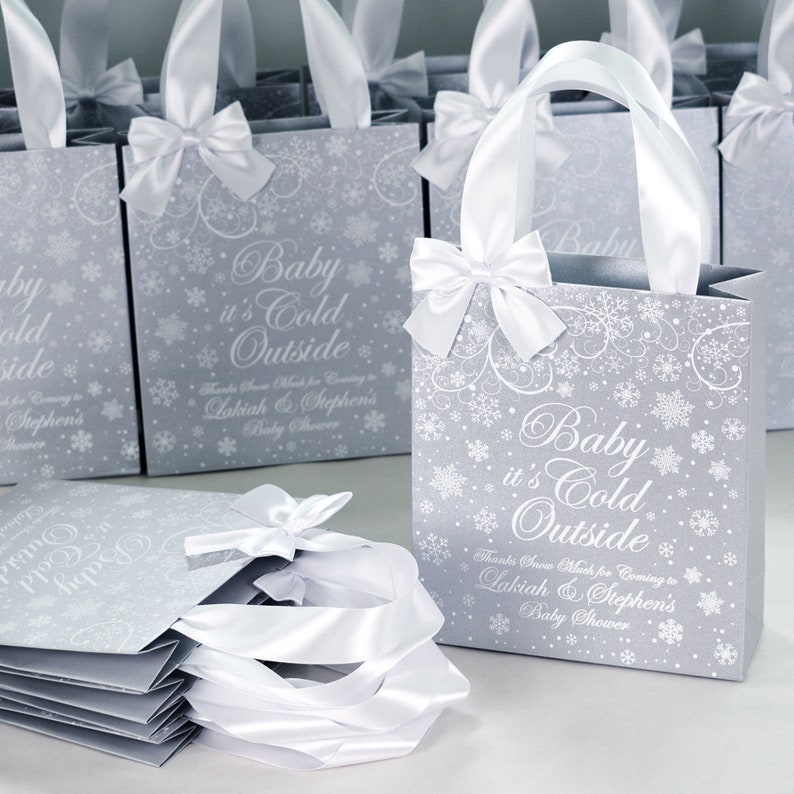 May include: Silver gift bags with white satin ribbon bows and snowflake designs. The bags have the text "Baby it's Cold Outside" and "Thanks Snow Much for Coming to Lakiah & Stephen's Baby Shower".