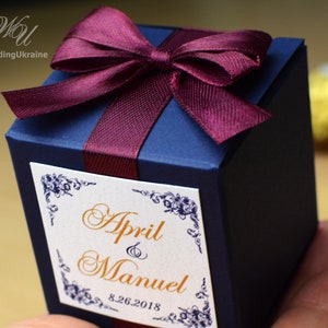 May include: A navy blue favor box with a burgundy satin ribbon tied in a bow. The box has a white label with gold lettering that reads "April & Manuel 8.26.2018".