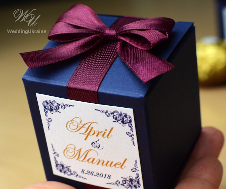 May include: A navy blue favor box with a burgundy satin ribbon tied in a bow. The box has a white label with gold lettering that reads "April & Manuel 8.26.2018".