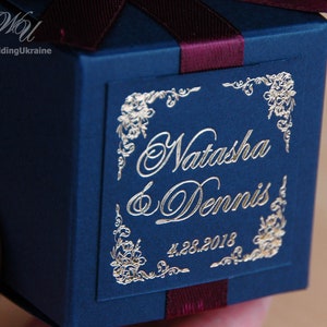 May include: A navy blue gift box tied with a burgundy ribbon. The box is adorned with gold foil lettering, reading "Natasha & Dennis" and the date "4.28.2018". Floral corner designs add a decorative touch.