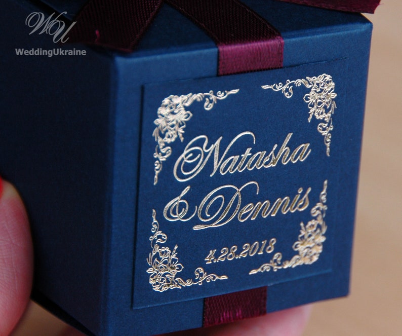 May include: A navy blue gift box tied with a burgundy ribbon. The box is adorned with gold foil lettering, reading "Natasha & Dennis" and the date "4.28.2018". Floral corner designs add a decorative touch.