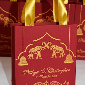 Op de afbeelding: Rode cadeauzakjes met gouden handvatten en gouden olifantmotieven. De zakjes hebben de tekst "Nithya & Christopher" en "30 december 2022".