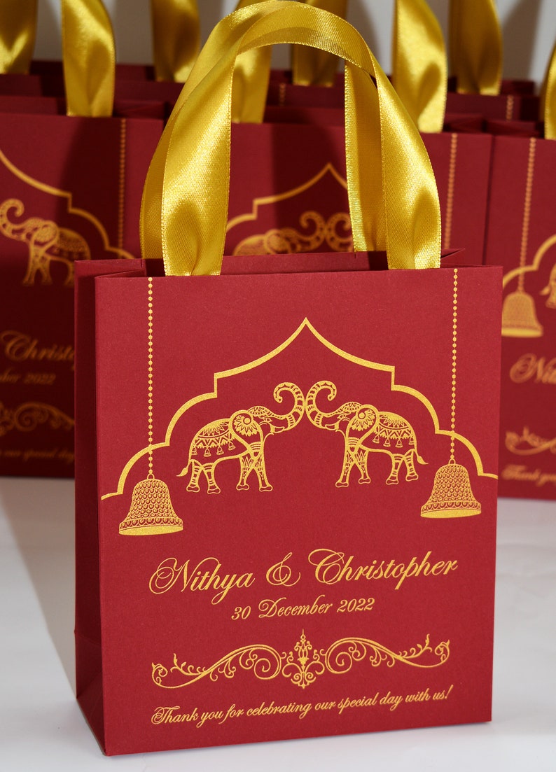 Op de afbeelding: Rode cadeauzakjes met gouden handvatten en gouden olifantmotieven. De zakjes hebben de tekst "Nithya & Christopher" en "30 december 2022".