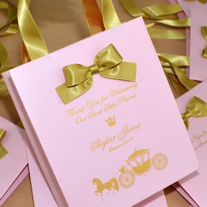May include: Pink favor bags with gold ribbon bows and a gold crown and carriage design. The bags have the text "Thank You for Welcoming Our Sweet Little Princess Skylar Jhene October 2019" printed on them.