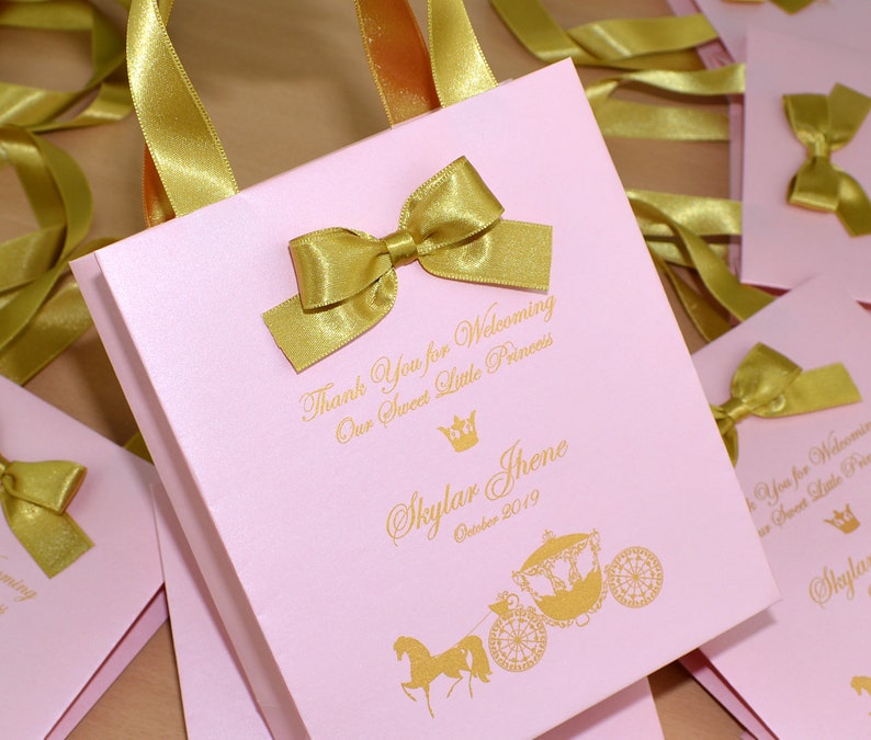 May include: Pink favor bags with gold ribbon bows and a gold crown and carriage design. The bags have the text "Thank You for Welcoming Our Sweet Little Princess Skylar Jhene October 2019" printed on them.