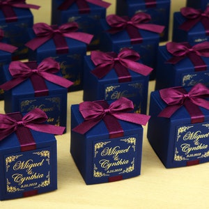 May include: Navy blue gift boxes tied with burgundy ribbon bows. Each box features a gold label with the names "Miguel & Cynthia" and the date "9.28.2019". The boxes are arranged on a light-coloured surface.