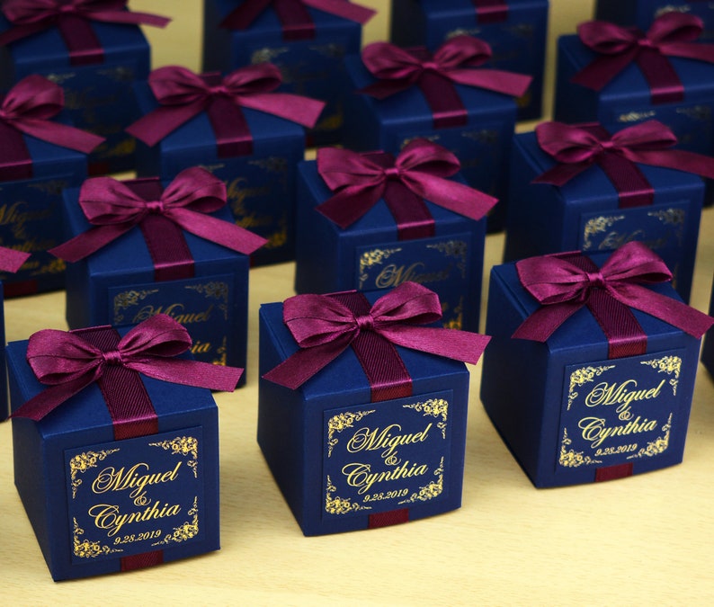 May include: Navy blue gift boxes tied with burgundy ribbon bows. Each box features a gold label with the names "Miguel & Cynthia" and the date "9.28.2019". The boxes are arranged on a light-coloured surface.
