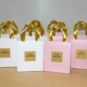 May include: A group of white and pink gift bags with gold ribbon handles. The bags are labeled with the text "Team Groom" and "Team Bride".