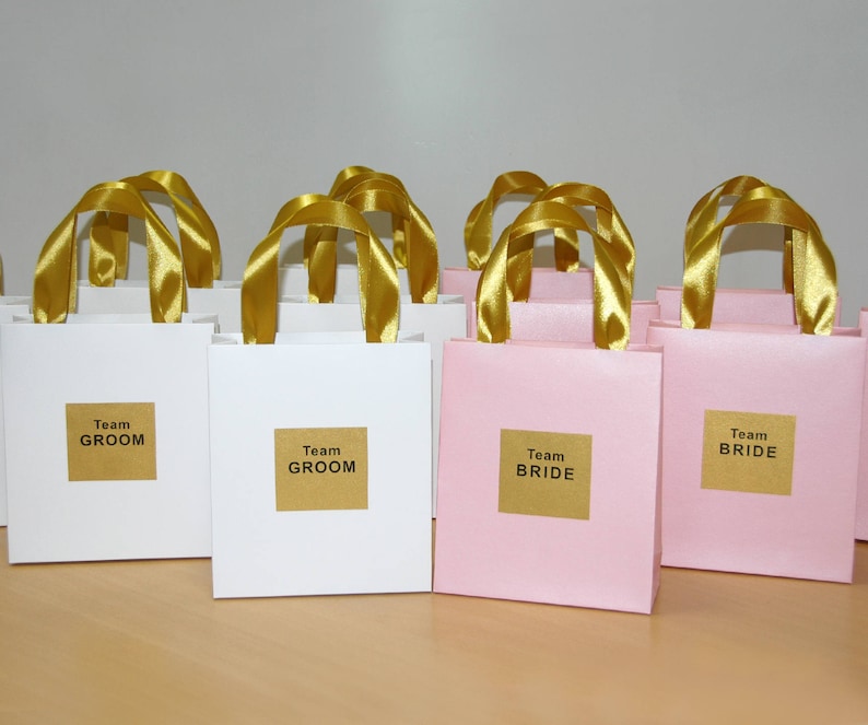 May include: A group of white and pink gift bags with gold ribbon handles. The bags are labeled with the text "Team Groom" and "Team Bride".