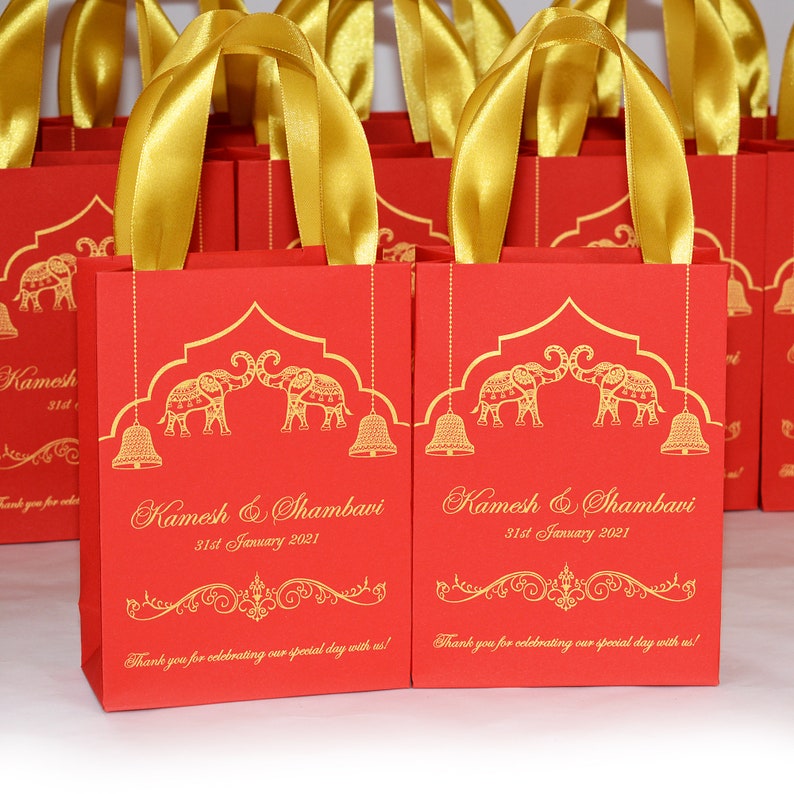 Op de afbeelding: Rode en gouden cadeauzakjes met een ontwerp van twee olifanten die elkaar aankijken. De zakjes hebben gouden handvatten en de tekst "Kamesh & Shambavi 31st January 2021" erop gedrukt.