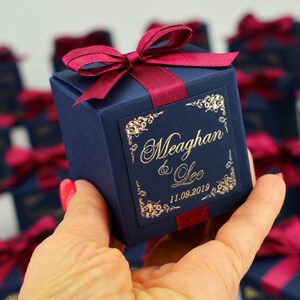 May include: A navy blue gift box tied with a burgundy ribbon and bow. The box is inscribed with gold lettering: "Meaghan & Lee" and the date "11.09.2019". Floral designs frame the text. Other similar boxes are visible in the background.