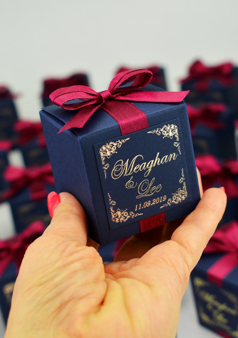 May include: A navy blue gift box tied with a burgundy ribbon and bow. The box is inscribed with gold lettering: "Meaghan & Lee" and the date "11.09.2019". Floral designs frame the text. Other similar boxes are visible in the background.