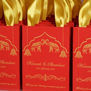 Op de afbeelding: Rode cadeauzakjes met gouden handvatten en gouden olifantmotieven. De zakken hebben de tekst "Kamesh & Shambavi 31st January 2021" en "Bedankt voor het vieren van onze speciale dag met ons!"