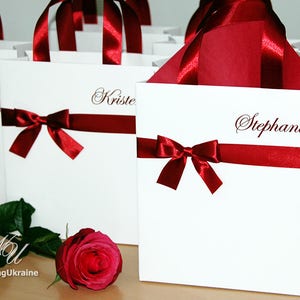 May include: White gift bags with red ribbon bows and personalized names, including "Maddie", "Krista", and "Stephanie".