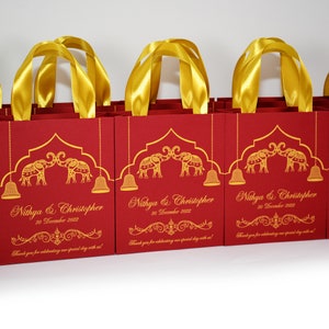 Op de afbeelding: Rode cadeauzakjes met gouden handvatten en gouden olifantmotieven. De zakjes hebben de tekst "Nithya & Christopher" en "30 december 2022" erop gedrukt. De zakjes hebben ook de tekst "Bedankt dat je onze speciale dag met ons hebt gevierd!" erop gedrukt.