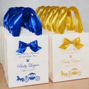 May include: White favor bags with blue and gold ribbon handles. The bags have a gold crown and carriage design with the text "Thank You for Welcoming Our Sweet Little Prince Baby Logan August 31st, 2019".