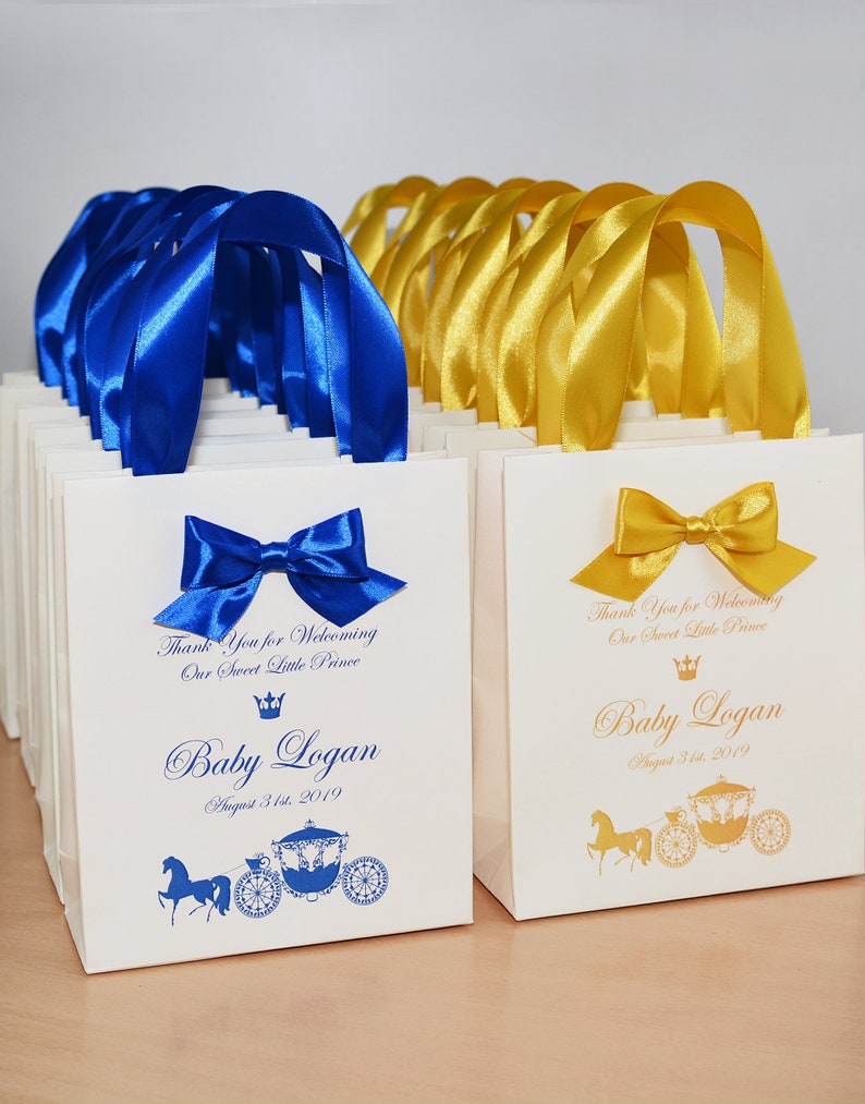 May include: White favor bags with blue and gold ribbon handles. The bags have a gold crown and carriage design with the text "Thank You for Welcoming Our Sweet Little Prince Baby Logan August 31st, 2019".