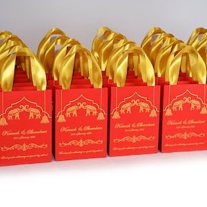 Op de afbeelding: Rode cadeautassen met gouden handvatten en gouden bedrukte olifanten, bellen en sierlijke ontwerpen. De tassen hebben de tekst "Kamesh & Shambavi 31st January 2021" erop gedrukt.