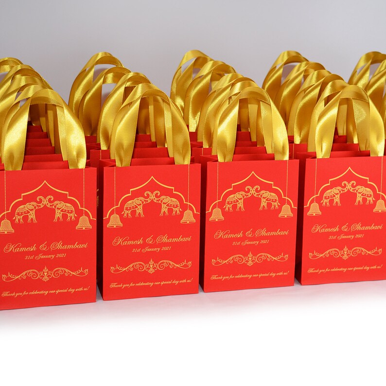 Op de afbeelding: Rode cadeautassen met gouden handvatten en gouden bedrukte olifanten, bellen en sierlijke ontwerpen. De tassen hebben de tekst "Kamesh & Shambavi 31st January 2021" erop gedrukt.