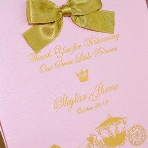 May include: Pink gift bag with a gold bow and gold lettering. The text reads "Thank You for Welcoming Our Sweet Little Princess Skylar Jhene October 2019". A gold crown and a gold carriage with a horse are also pictured.