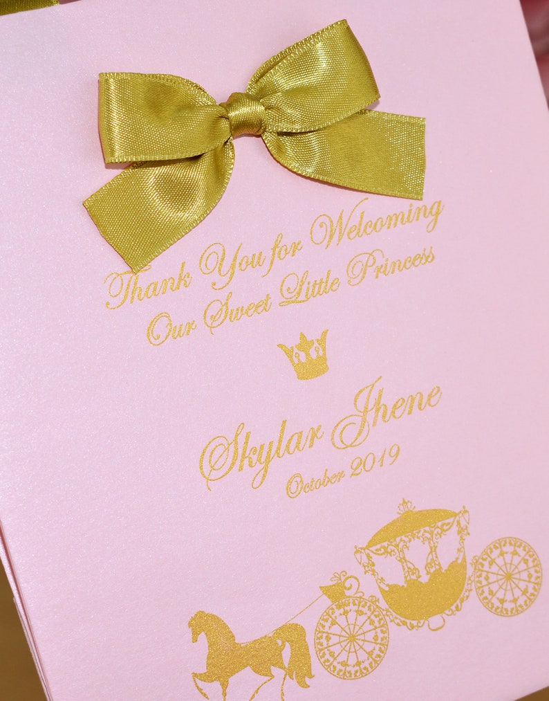 May include: Pink gift bag with a gold bow and gold lettering. The text reads "Thank You for Welcoming Our Sweet Little Princess Skylar Jhene October 2019". A gold crown and a gold carriage with a horse are also pictured.