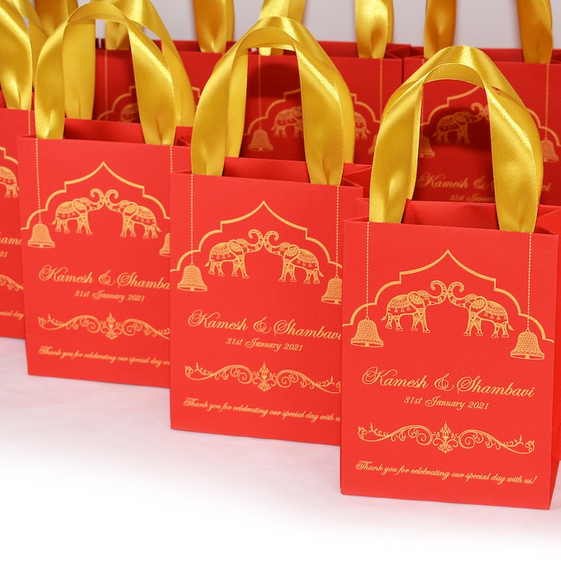 Op de afbeelding: Rode cadeauzakjes met gouden handvatten en gouden olifantmotieven. De zakjes hebben de tekst "Kamesh & Shambavi 31st January 2021" erop gedrukt. Bedankt dat je onze speciale dag met ons hebt gevierd!