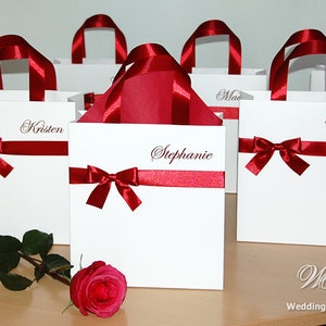 May include: White gift bags with red ribbon bows and personalized names, including Kristen, Stephanie, and Catey. The bags are arranged on a wooden surface with a single red rose.