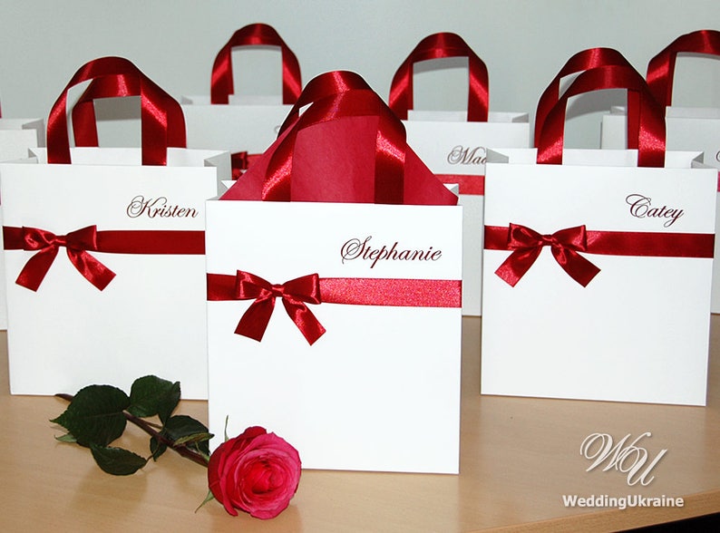 May include: White gift bags with red ribbon bows and personalized names, including Kristen, Stephanie, and Catey. The bags are arranged on a wooden surface with a single red rose.