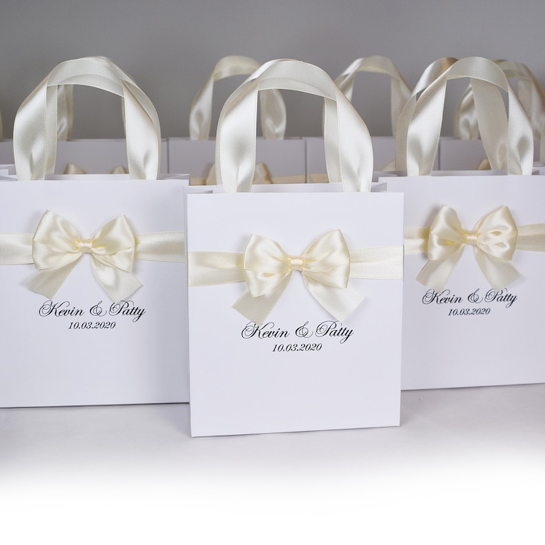 Puede incluir: Bolsas de regalo blancas con lazos de cinta de sat&eacute;n marfil y el texto "Kevin & Patty 10.03.2020" impreso en la parte delantera.