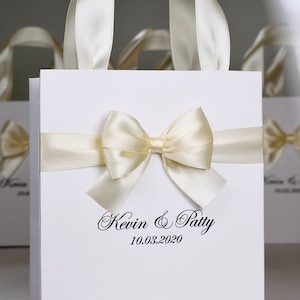 Puede incluir: Bolsa de regalo blanca con una cinta de sat&eacute;n y un lazo. La bolsa tiene el texto "Kevin & Patty 10.03.2020" impreso en la parte delantera.
