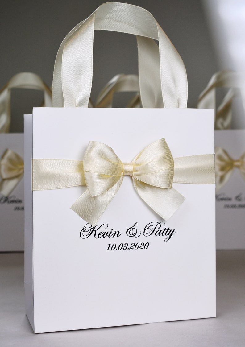 Puede incluir: Bolsa de regalo blanca con una cinta de sat&eacute;n y un lazo. La bolsa tiene el texto "Kevin & Patty 10.03.2020" impreso en la parte delantera.